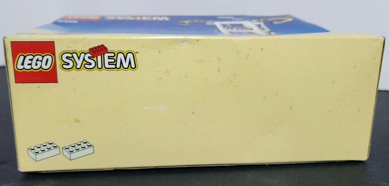 Puede incluir: Una caja blanca con el logotipo de LEGO SYSTEM en rojo y azul. La caja tiene un fondo azul con nubes blancas. La caja es para un set de LEGO.