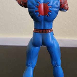May include: A Spider-Man action figure, viewed from the back, in a dynamic pose. The figure is predominantly blue with red accents, including the head, hands, and feet. A red spider emblem is on the back. The figure is on a dark surface.