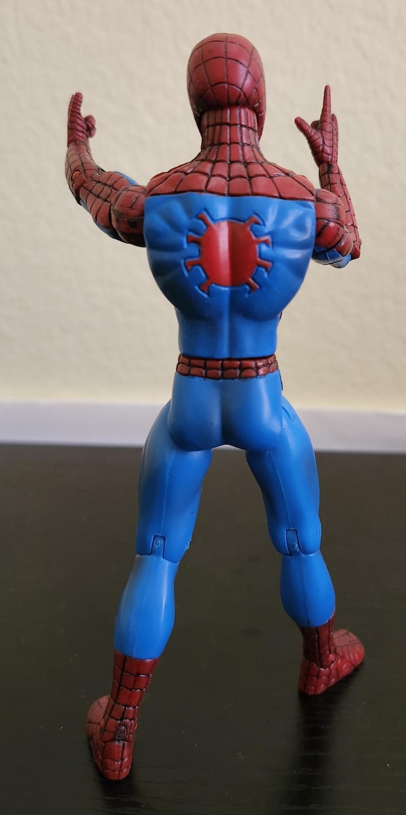 May include: A Spider-Man action figure, viewed from the back, in a dynamic pose. The figure is predominantly blue with red accents, including the head, hands, and feet. A red spider emblem is on the back. The figure is on a dark surface.