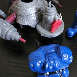 May include: A selection of plastic toy components, featuring a silver spider-shaped piece, grey cylindrical parts with red tips, and a blue torso. These elements suggest a larger action figure or toy set, disassembled for play or storage.