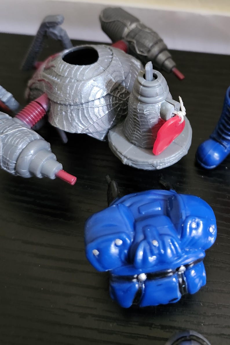 May include: A selection of plastic toy components, featuring a silver spider-shaped piece, grey cylindrical parts with red tips, and a blue torso. These elements suggest a larger action figure or toy set, disassembled for play or storage.