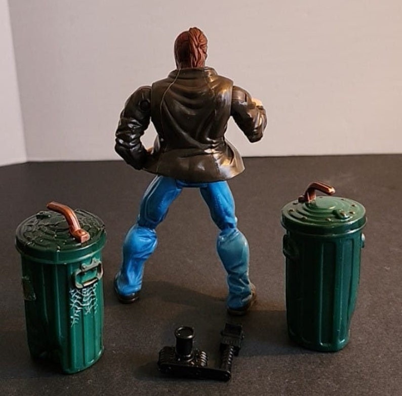 May include: A brown-haired action figure wearing a brown leather jacket, blue jeans, and black boots. The figure is holding a small black object in its right hand. The figure is standing in front of two green trash cans with brown lids and handles.