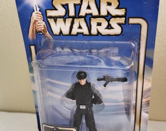 May include: A Star Wars action figure of an Imperial Officer from the movie 'A New Hope'. The figure is wearing a black uniform with a black cap and is holding a blaster.