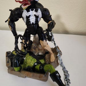 May include: A black and white Venom action figure with a white spider emblem on its chest, battling a green figure. The Venom figure has a large mouth with a red tongue and is posed in a dynamic action stance. The figures are on a detailed base.