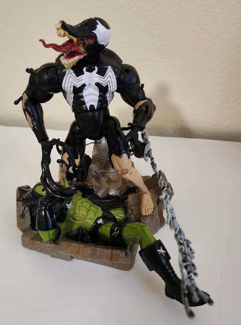 May include: A black and white Venom action figure with a white spider emblem on its chest, battling a green figure. The Venom figure has a large mouth with a red tongue and is posed in a dynamic action stance. The figures are on a detailed base.