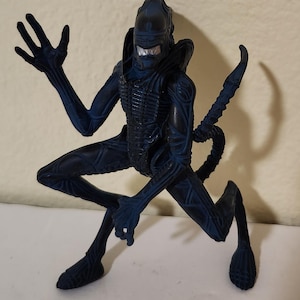 Puede incluir: Figura de acción Xenomorfo azul oscuro en posición agachada. La figura tiene una cola larga y segmentada y un exoesqueleto detallado. Los brazos de la criatura están extendidos y su cabeza presenta un cráneo liso y alargado.