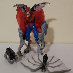 May include: A Spider-Man action figure with a red and blue costume, red cape, and bat-like wings. The figure has gray, monster-like legs and arms. Additional accessories include a black claw and a white and gray spider-wing accessory.