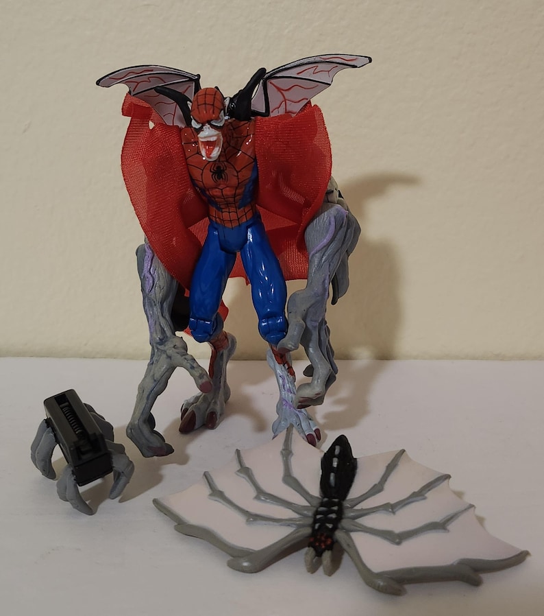 May include: A Spider-Man action figure with a red and blue costume, red cape, and bat-like wings. The figure has gray, monster-like legs and arms. Additional accessories include a black claw and a white and gray spider-wing accessory.