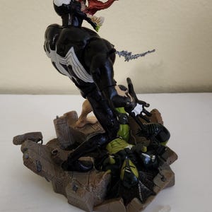 May include: A detailed action figure of Venom, a Marvel Comics character, in a dynamic pose. The figure is predominantly black with white accents and a large, open mouth displaying a red tongue and teeth. The base is a detailed, textured brown. A clear plastic stand is included.
