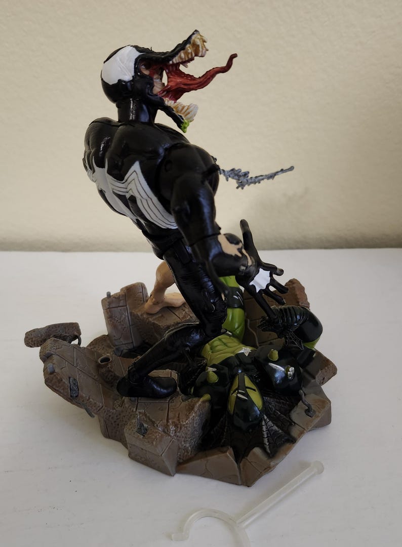 May include: A detailed action figure of Venom, a Marvel Comics character, in a dynamic pose. The figure is predominantly black with white accents and a large, open mouth displaying a red tongue and teeth. The base is a detailed, textured brown. A clear plastic stand is included.