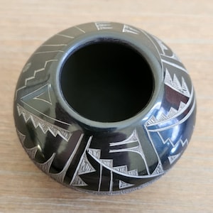 May include: A black, round ceramic pot with a wide opening. The pot features intricate, etched geometric designs in white around the exterior. The pot has a glossy finish and sits on a light-coloured surface.