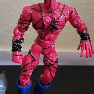 May include: A red action figure of a superhero with black web-like detailing and blue boots. The figure has a spider emblem on the chest and black detailing around the arms. The figure is posed with a dynamic stance.