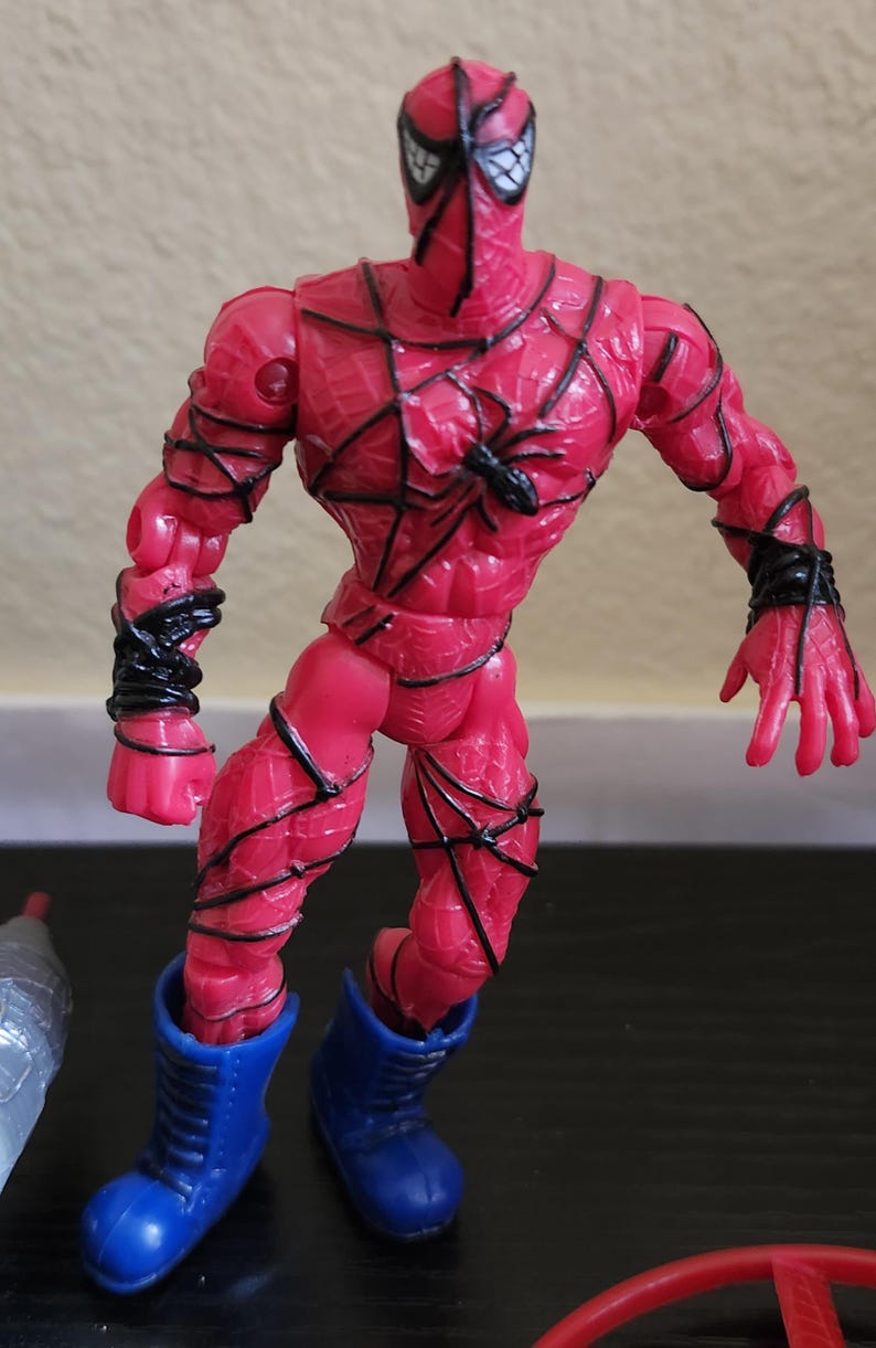 May include: A red action figure of a superhero with black web-like detailing and blue boots. The figure has a spider emblem on the chest and black detailing around the arms. The figure is posed with a dynamic stance.