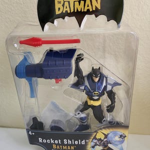 May include: A blue and black Batman action figure with a rocket shield accessory. The figure is packaged in a clear plastic blister pack with a red and blue background. The packaging features the text "The Batman" and "Rocket Shield".