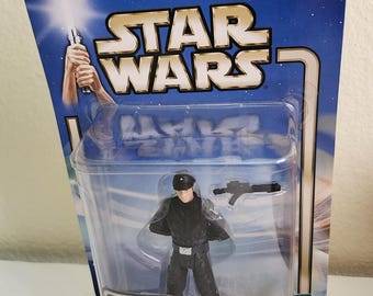 May include: A Star Wars action figure of an Imperial Officer from the movie A New Hope. The figure is wearing a black uniform with a black helmet and holding a blaster.