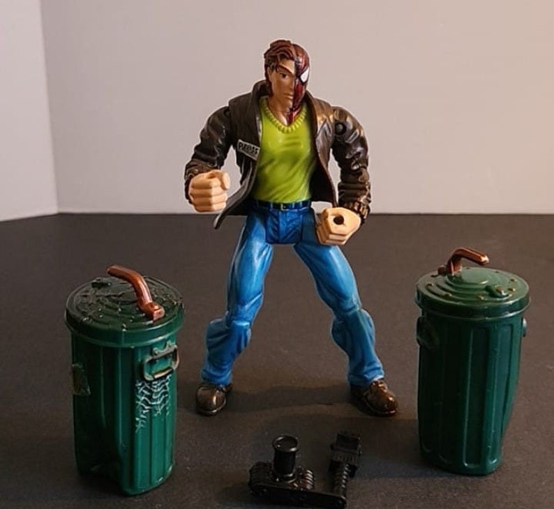 May include: A green and brown plastic toy figure of a man with half of his face painted red and the other half painted brown. The figure is wearing a green shirt, blue jeans, and a brown leather jacket. The figure is standing in a fighting pose with his fists clenched. The figure is surrounded by two green plastic toy trash cans.