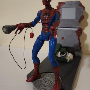 以下が含まれることがあります： 赤と青のクラシックなスーツを着たスパイダーマンのアクションフィギュア。ウェブシューターデバイスを持っています。フィギュアは円形のデザインの灰色のプラットフォームに立っており、近くにスパイダーマンの頭部が分離しています。背景には壊れた壁があります。