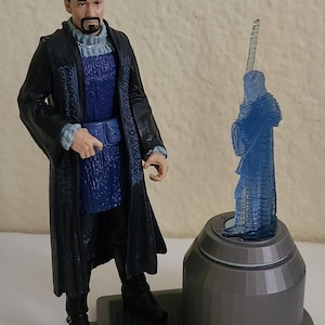 May include: A detailed action figure of a character in a long black coat with blue accents, standing next to a blue translucent figure on a gray base. The figure is wearing black boots.