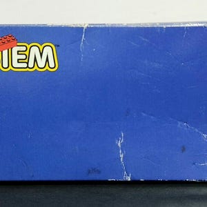 Puede incluir: Una caja azul LEGO System con el logotipo de LEGO y un ladrillo LEGO rojo en la parte delantera.