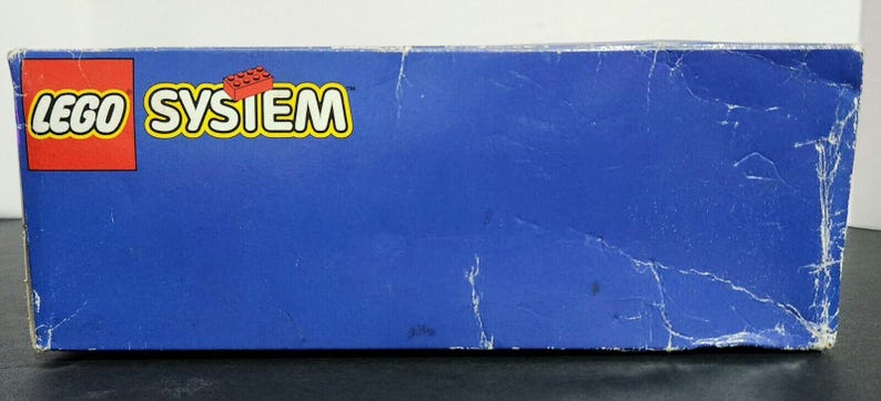 Puede incluir: Una caja azul LEGO System con el logotipo de LEGO y un ladrillo LEGO rojo en la parte delantera.