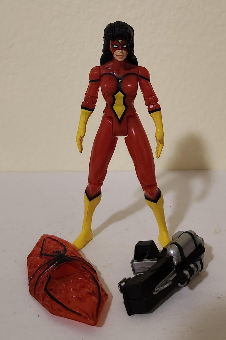 May include: A red and yellow action figure of a female superhero, with black hair and a mask. The figure is standing with accessories including a red shield with a black spider design and a black and silver weapon.