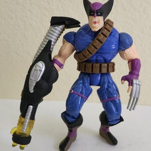 May include: Action figure of a superhero in a blue suit with a brown belt and purple accents. The figure has a mask, black headgear, and a weapon. The figure has a stern expression and metal claws.