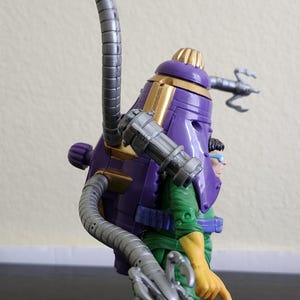 May include: A purple and gold action figure with a large, cylindrical head and multiple mechanical arms. The figure is wearing a green and yellow outfit. The figure has a unique design with a futuristic aesthetic.