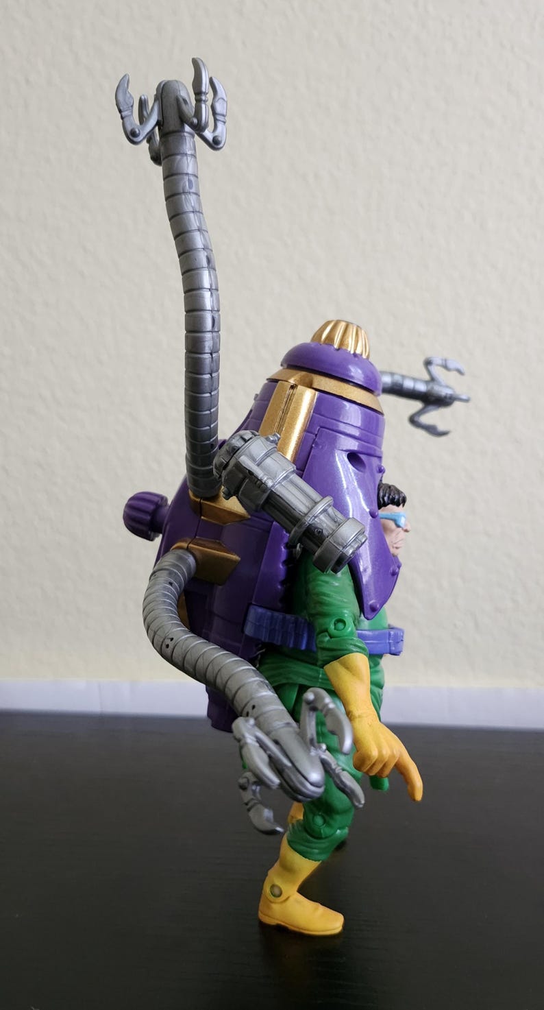 May include: A purple and gold action figure with a large, cylindrical head and multiple mechanical arms. The figure is wearing a green and yellow outfit. The figure has a unique design with a futuristic aesthetic.