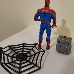 May include: A Spider-Man action figure in a red and blue suit, with a black spider web and a gray device. The figure is standing, and the accessories are placed nearby. The toy is a collectible item.