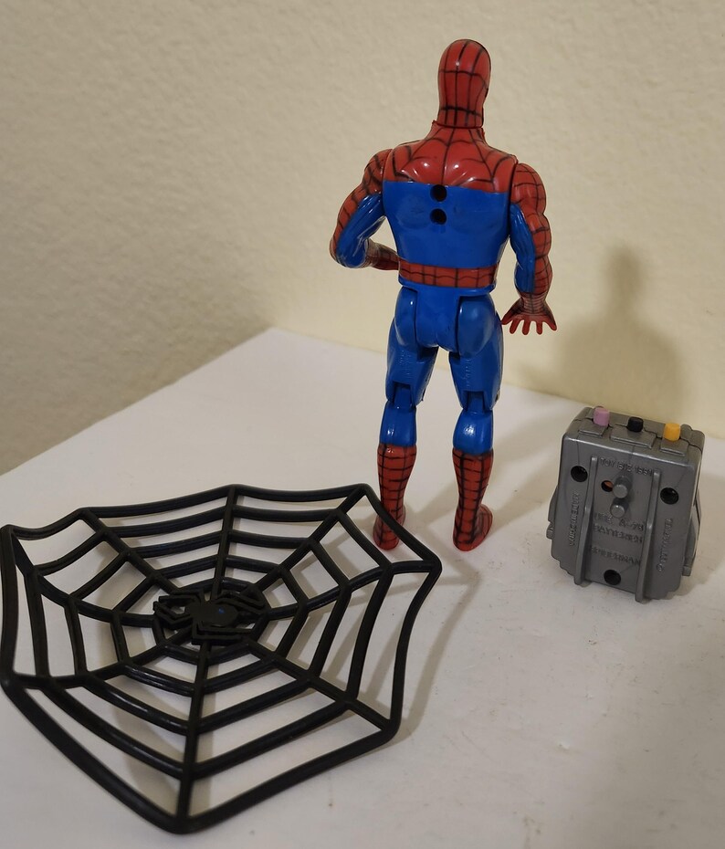 May include: A Spider-Man action figure in a red and blue suit, with a black spider web and a gray device. The figure is standing, and the accessories are placed nearby. The toy is a collectible item.