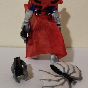 May include: A detailed action figure with a red cape, gray arms, and a black headpiece. The figure has detached wings and a separate gray hand holding a black object. The figure is on a white surface.