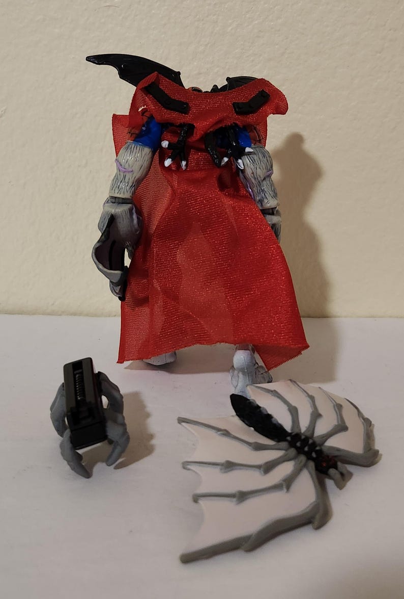 May include: A detailed action figure with a red cape, gray arms, and a black headpiece. The figure has detached wings and a separate gray hand holding a black object. The figure is on a white surface.