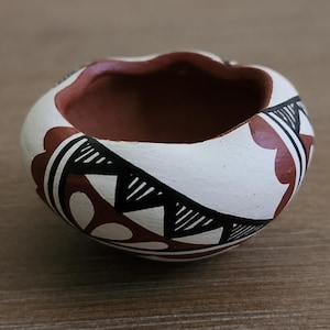 Puede incluir: Un pequeño cuenco de cerámica pintado a mano con una forma irregular. El exterior es blanco con patrones geométricos marrones y negros. El interior es marrón, creando un contraste. Aproximadamente 10 cm de diámetro.