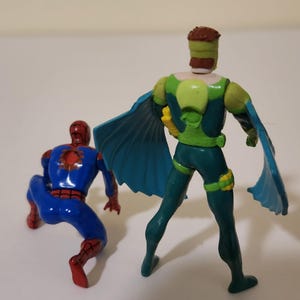 Pu&ograve; includere: Due action figure di supereroi. Una figura &egrave; accovacciata, indossa un costume blu e rosso. L'altra figura &egrave; in piedi, indossa un costume verde e blu con un mantello. Le figure sono su una superficie bianca.