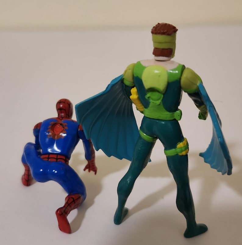 Pu&ograve; includere: Due action figure di supereroi. Una figura &egrave; accovacciata, indossa un costume blu e rosso. L'altra figura &egrave; in piedi, indossa un costume verde e blu con un mantello. Le figure sono su una superficie bianca.