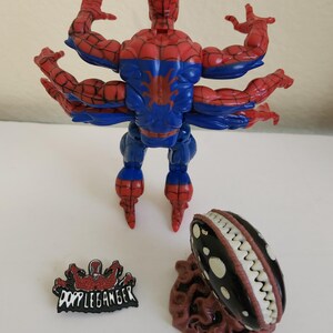 May include: A Spider-Man action figure with six arms, in red and blue, and a black and white creature with teeth. Also included is a "Doppleganger" pin.