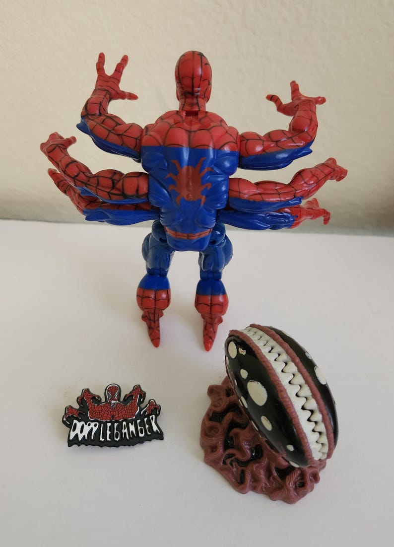 May include: A Spider-Man action figure with six arms, in red and blue, and a black and white creature with teeth. Also included is a "Doppleganger" pin.