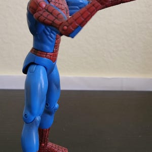 May include: A Spider-Man action figure in a dynamic pose, featuring a red and blue costume. The figure has a red mask with white eye lenses and is striking a hand gesture. The figure is approximately 6 inches tall.