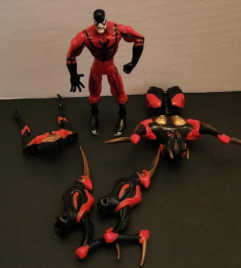 May include: A red and black action figure with a black mask and a spider symbol on its chest. The figure is standing on a white surface. There are several other pieces of the figure, including a pair of legs, a torso, and a head. The pieces are all black and red.