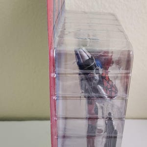 May include: A red, white and blue action figure in a plastic blister pack. The figure is holding a weapon and is posed in a dynamic action pose.