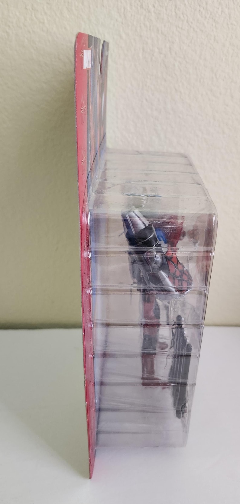May include: A red, white and blue action figure in a plastic blister pack. The figure is holding a weapon and is posed in a dynamic action pose.