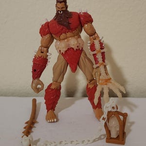 Może przedstawiać: Figurka akcji muskularnego, ubranego na czerwono wojownika z brązową brodą i kolczastą zbroją. Figurka ma przepaskę biodrową, akcenty w kształcie czaszki i akcesoria, w tym maczugę, łańcuch i klatkę z czaszką.