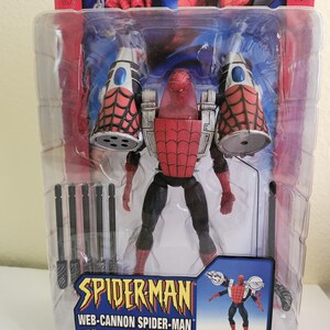 May include: A red and black action figure of Spider-Man wearing a web-cannon suit. The figure is packaged in a plastic blister pack with a blue and white background. The packaging includes the text "SPIDER-MAN WEB-CANNON SPIDER-MAN with Missile Launcher & Grappling Hook Action!"
