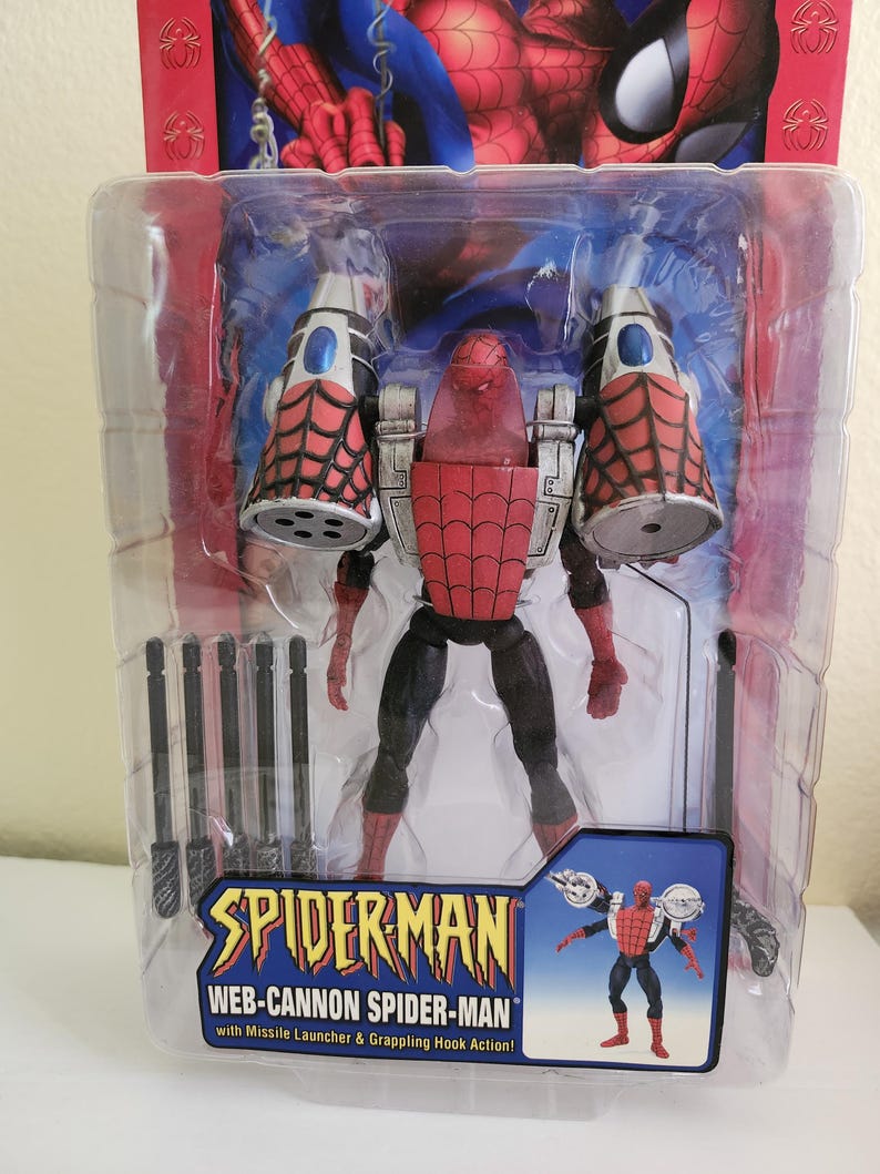 May include: A red and black action figure of Spider-Man wearing a web-cannon suit. The figure is packaged in a plastic blister pack with a blue and white background. The packaging includes the text "SPIDER-MAN WEB-CANNON SPIDER-MAN with Missile Launcher & Grappling Hook Action!"