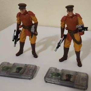 Könnte beinhalten: Zwei Actionfiguren eines Soldaten in brauner Uniform und Mütze, die eine Waffe halten. Sie stehen neben zwei transparenten Kunststoffboxen mit kleinen Bildern. Die Uniformen der Soldaten sind orange und gelb.