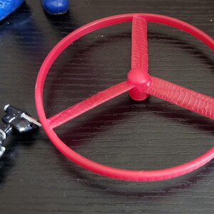May include: A red plastic toy propeller with three blades and a circular frame. The propeller is positioned on a dark surface, with a small, articulated black and silver toy component nearby. Blue toy shoes are visible in the background.