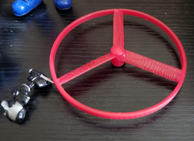 May include: A red plastic toy propeller with three blades and a circular frame. The propeller is positioned on a dark surface, with a small, articulated black and silver toy component nearby. Blue toy shoes are visible in the background.