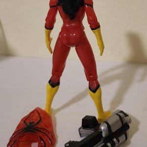 May include: A Spider-Woman action figure, viewed from the back, in a red and yellow costume. The figure has black hair and is standing with a web-like accessory and a silver and black device. The figure is approximately 6 inches tall.