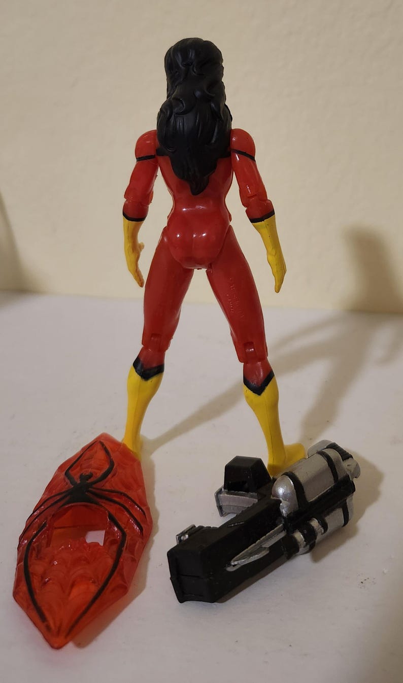 May include: A Spider-Woman action figure, viewed from the back, in a red and yellow costume. The figure has black hair and is standing with a web-like accessory and a silver and black device. The figure is approximately 6 inches tall.