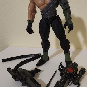 Può includere: Action figure di un personaggio muscoloso con maschera, pantaloni neri e armatura verde e nera. Gli accessori includono una pistola, una lancia con punta rossa e altre parti di armi. La figura è in piedi.